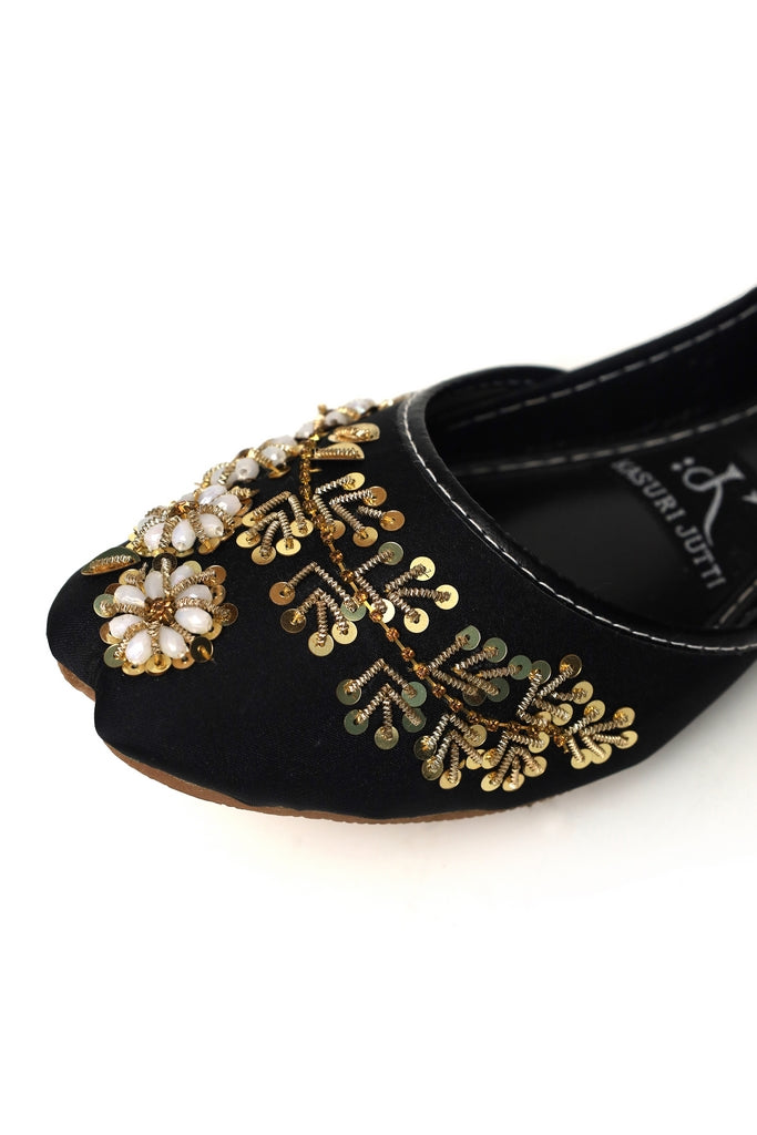 Black kasuri jutti with gold and pearl embellishments on a white background, closeup